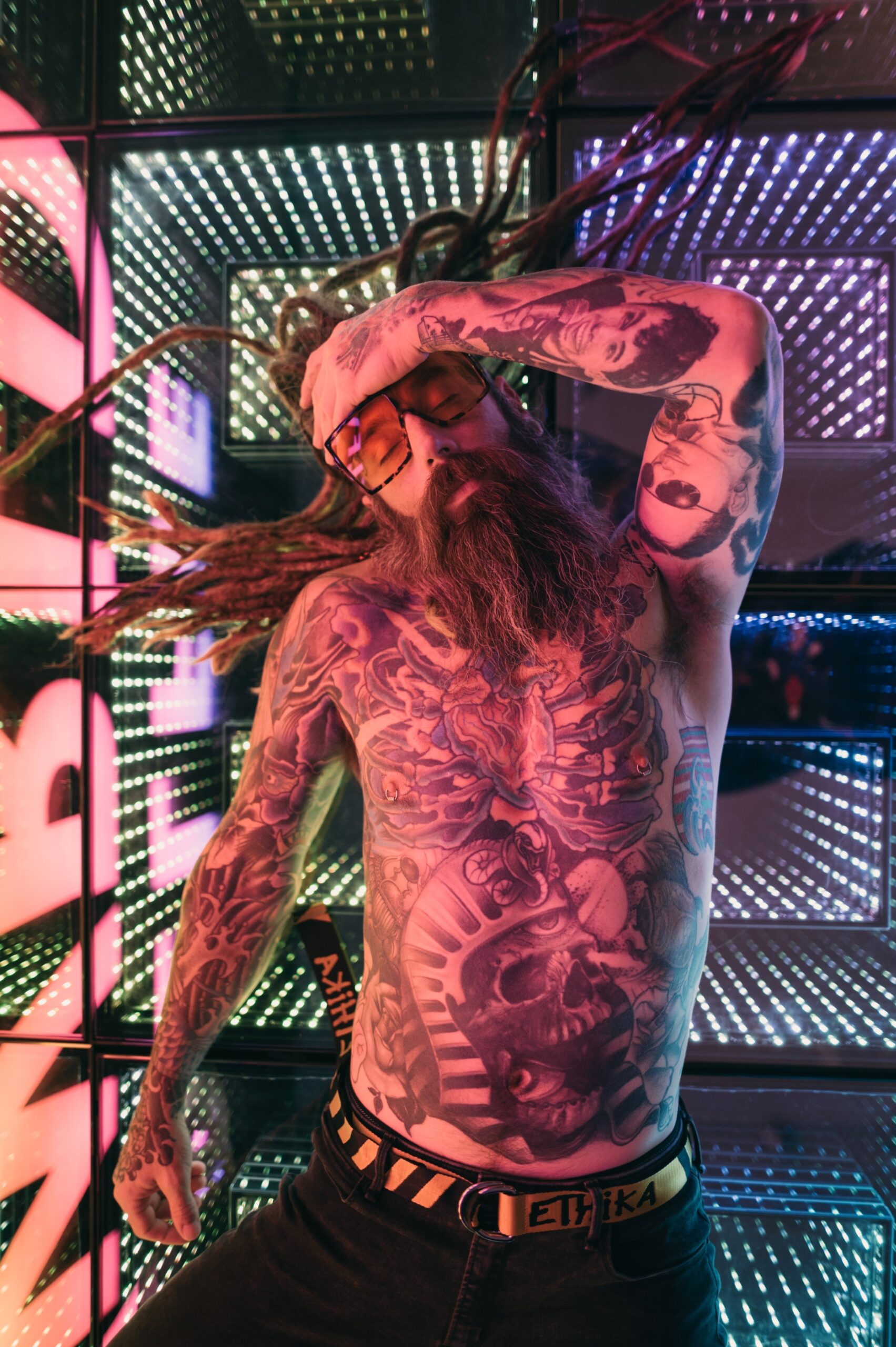 Shirtless tattooed man with dreadlocks, a beard, and sunglasses laying on glass floor with vibrant LED lights in it.