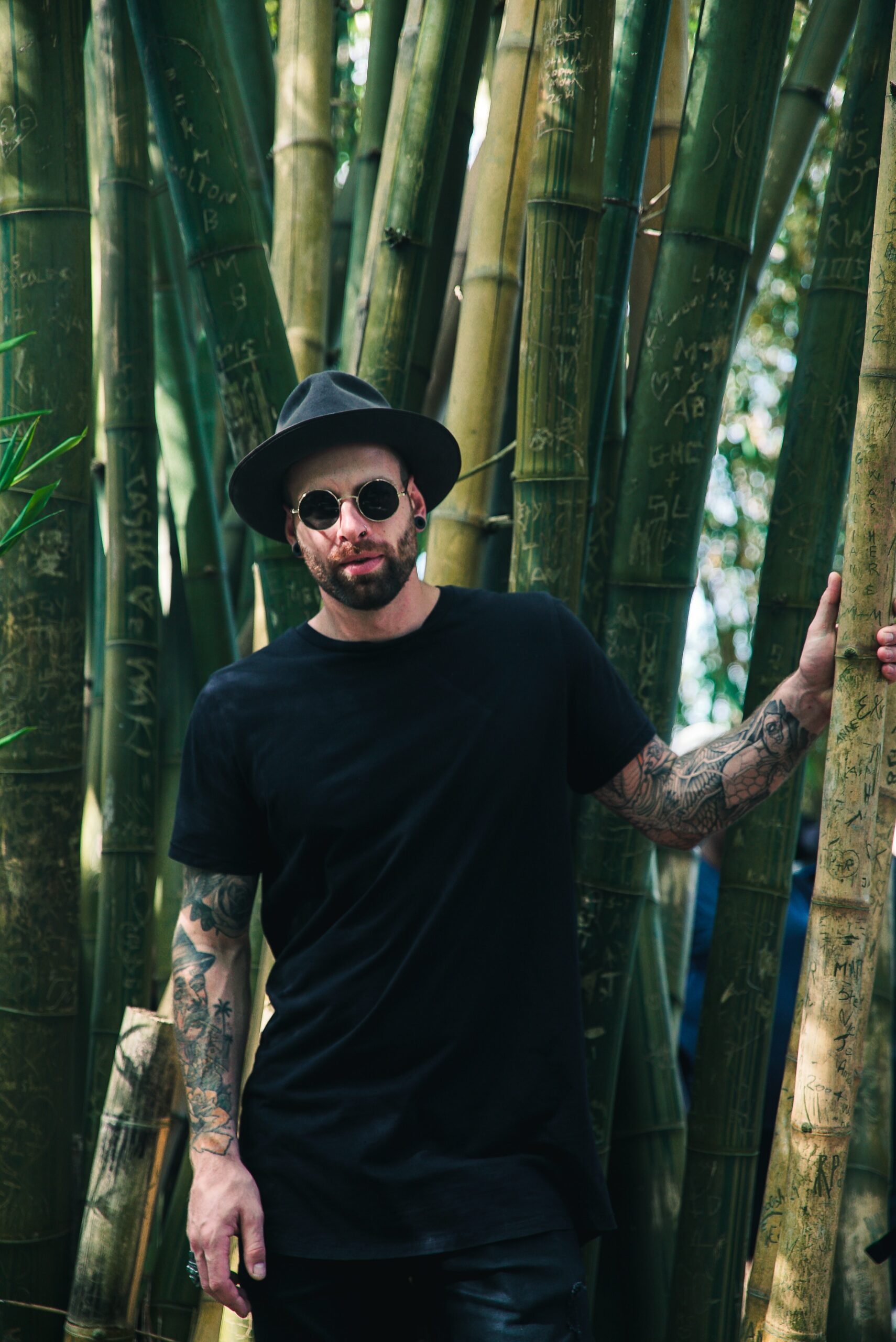 Man in all black with bucket hat wearing black sunglasses standing in the woods with full arm tattoos.