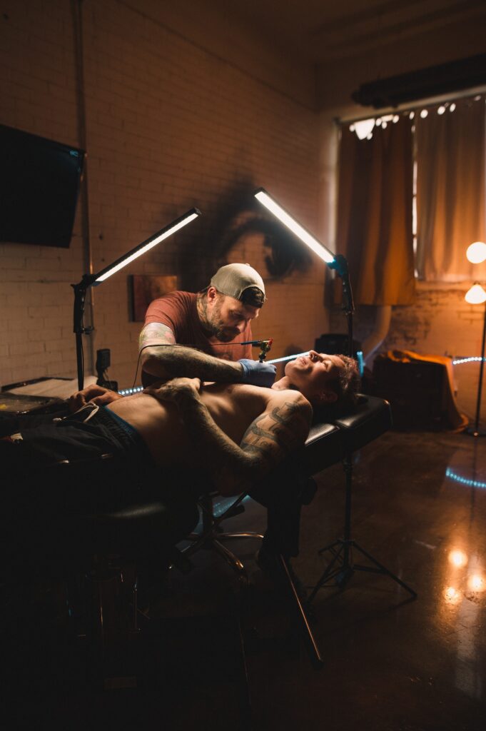 Man tattooing males chest in ambient lit tattoo studio.