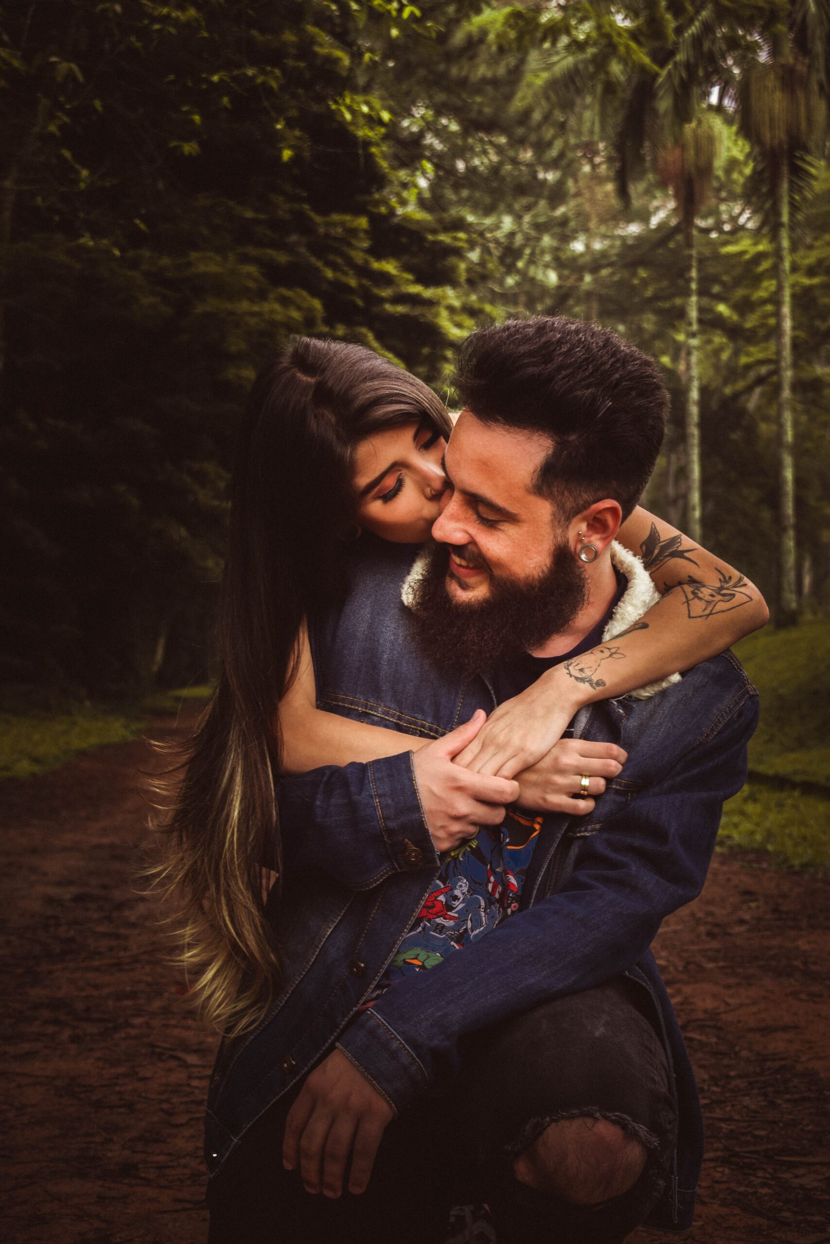 Girl with long black hair holding man with beard and jacket from behind kissing him on the cheek. They are standing on a dirt road in the middle of the woods.