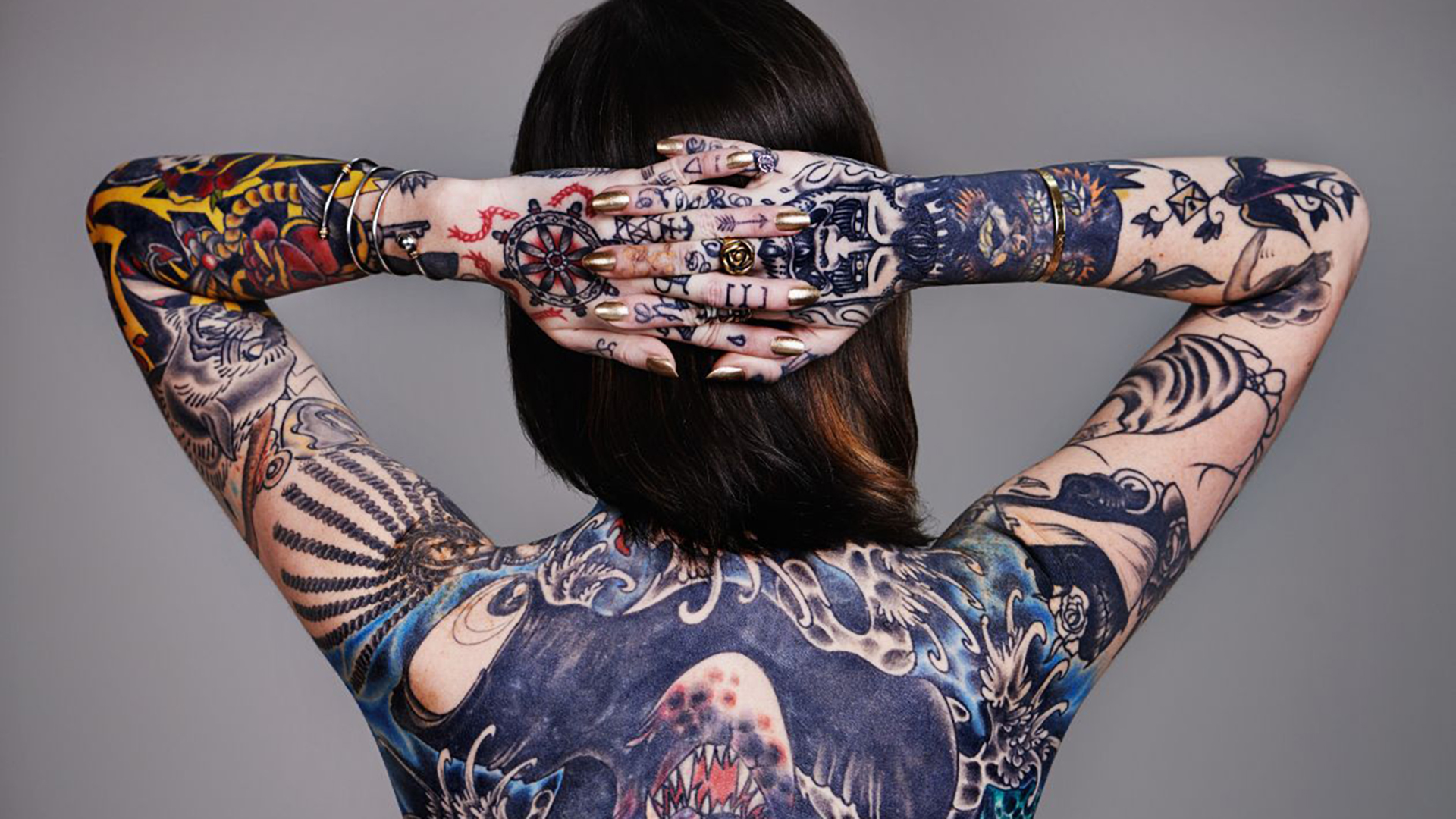 Woman photographed from behind with full back and arm tattoo coverage, hands clasped behind her head